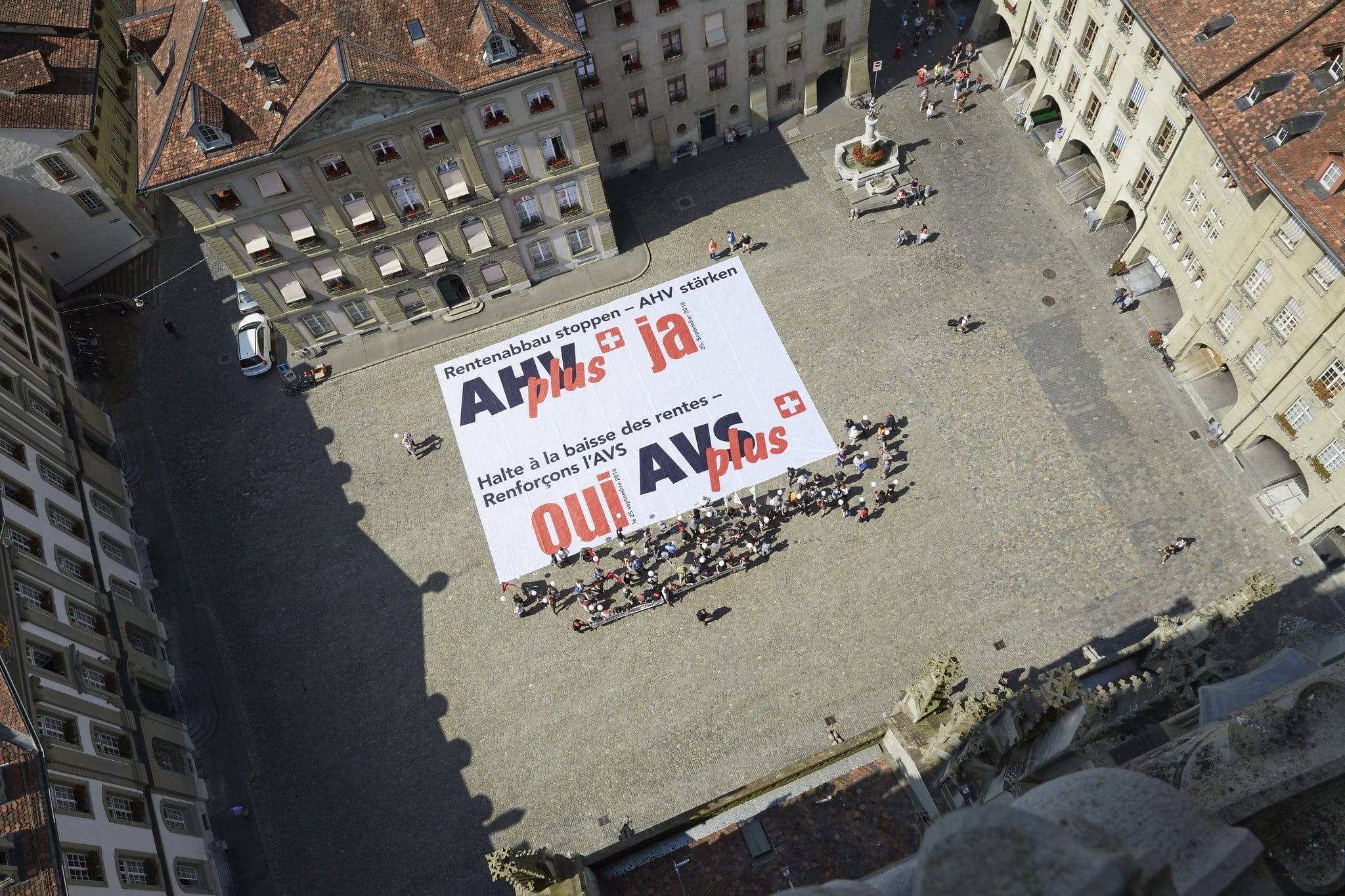 AHV: Wofür steht das «V»?Mitglieder des SGB entrollen ein Transparent zur AHV-Plus-Initiative auf dem Muensterplatz in Bern. Bild: Yoshiko Kusano auf flickr.com.